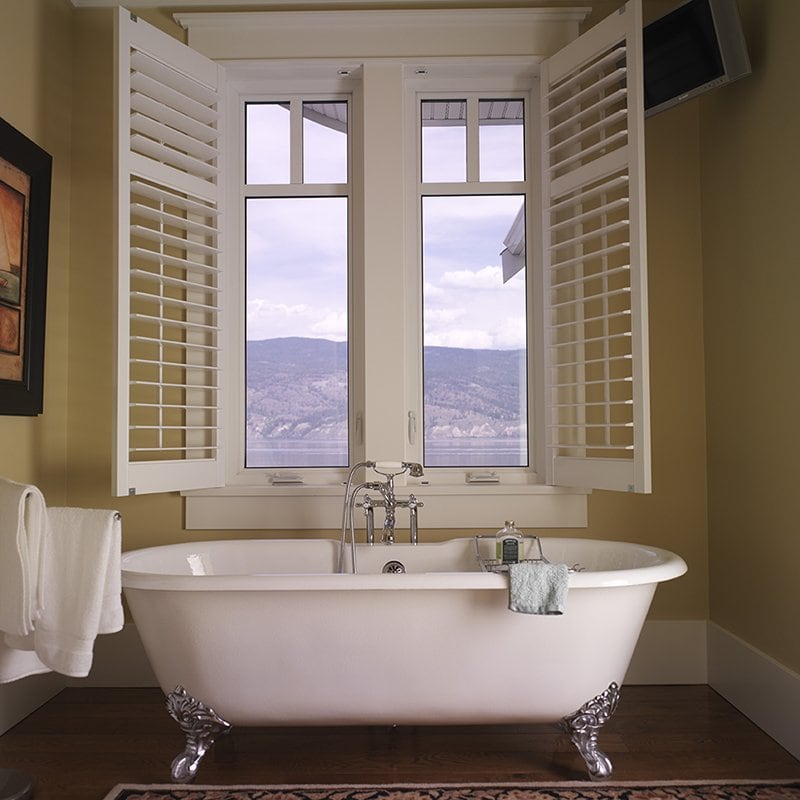 A claw-foot bathtub with a window above it with the window’s shutters opened into the bathroom.